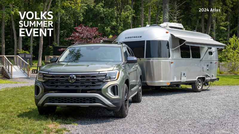An Atlas Peak Edition in Avocado Green Pearl is parked on gravel with grassy surroundings. In the background, is a forested area with tall trees. Behind the Atlas, is a chrome trailer which is hitched onto the back of the SUV.
