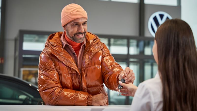 Ein Mann in orangener Jacke gibt Autoschlüssel beim Händler ab