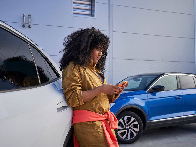 Nainen nojaa Volkswageniin ja katsoo matkapuhelintaan