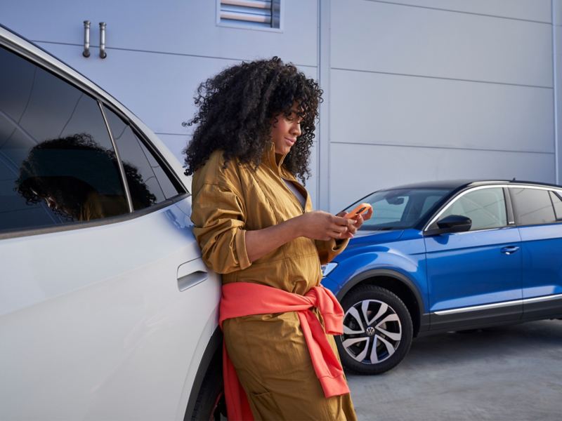 Nainen nojaa Volkswageniin ja katsoo matkapuhelintaan