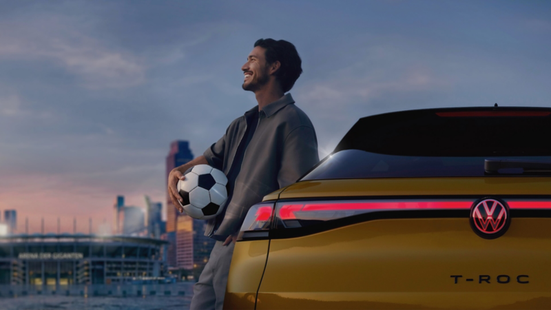 Mann mit Fußball lehnt an einem gelben VW T‑Roc vor einer Stadionkulisse bei Sonnenuntergang​