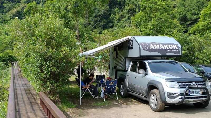 改裝後的銀黑雙色Amarok停在露營區，加裝的側帳拉開後，車主的家人在側帳下方的露營桌椅聊天休息