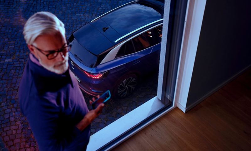 Een man kijkt op zijn smartphone, op de achtergrond zijn de achterkant en het dak van een blauwe VW ID.4 te zien door een raam.