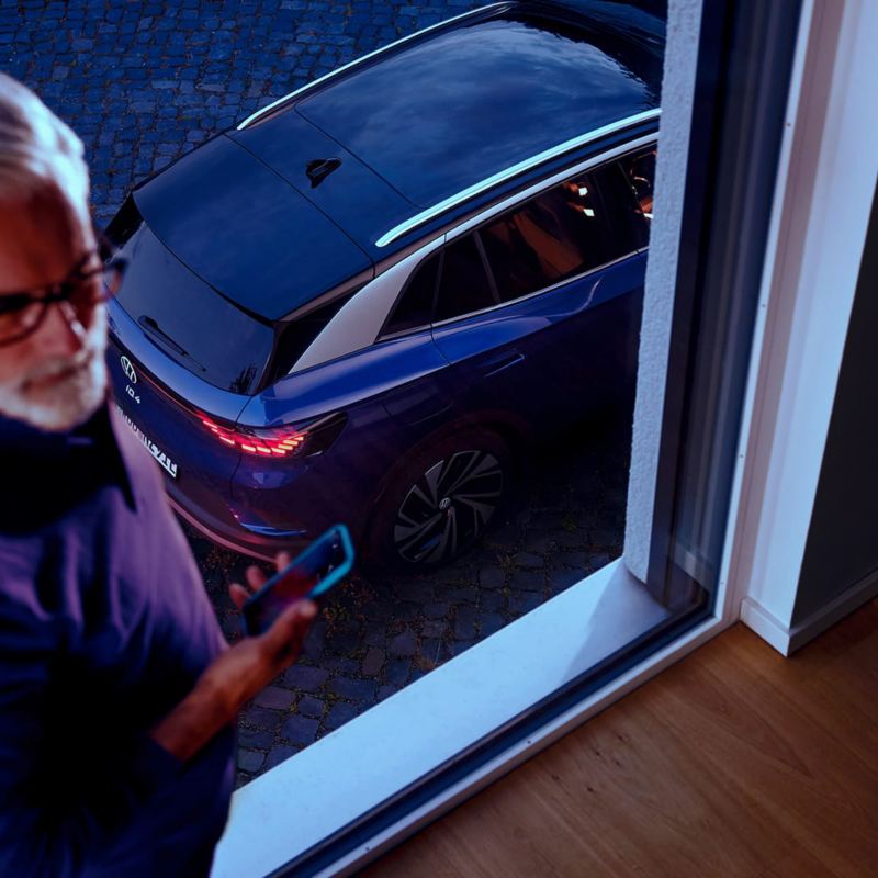 Un homme regarde son smartphone, à l’arrière-plan, une fenêtre à travers de laquelle on aperçoit le hayon et le toit d’une VW ID.4 bleue