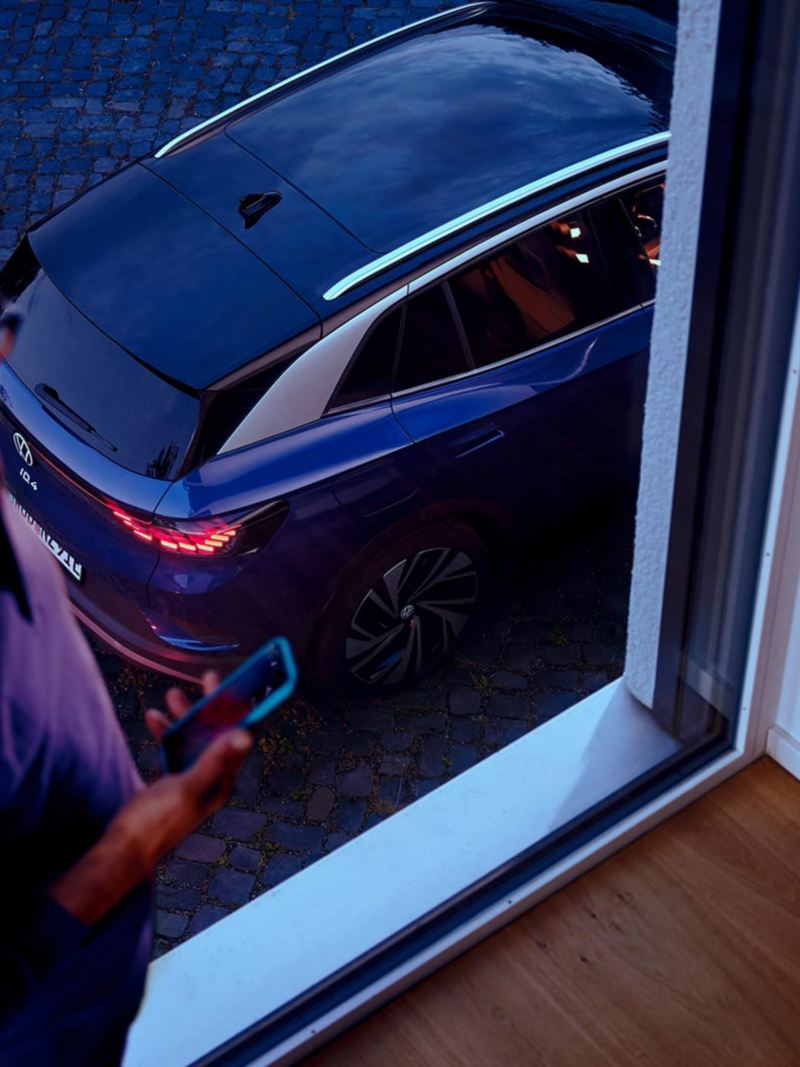 Un homme regarde son smartphone, à l’arrière-plan, une fenêtre à travers de laquelle on aperçoit le hayon et le toit d’une VW ID.4 bleue