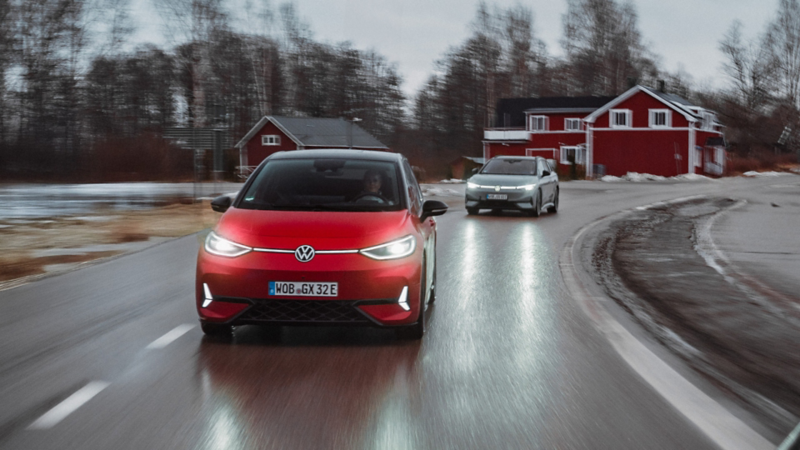 Ein roter ID.3 GTX fährt auf einer Landstraße vor einem grauen ID.7. Im Hintergrund sind rote Holzhäuser.