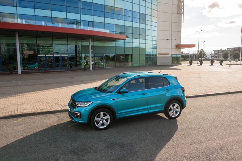 VW T-Cross R-Line stands in front of a beach and sea, a woman dressed in summer clothes gets out of the driver’s side.