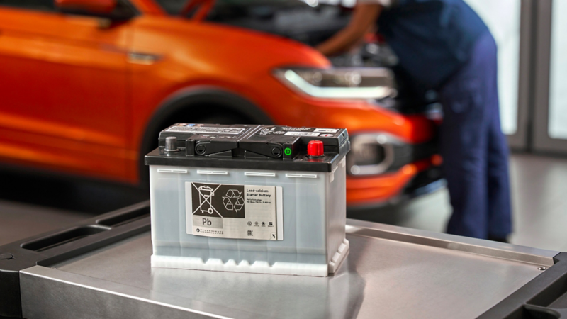 Volkswagen Genuine Lead-Calcium Starter Battery on a roller container, a VW T-Cross in the background