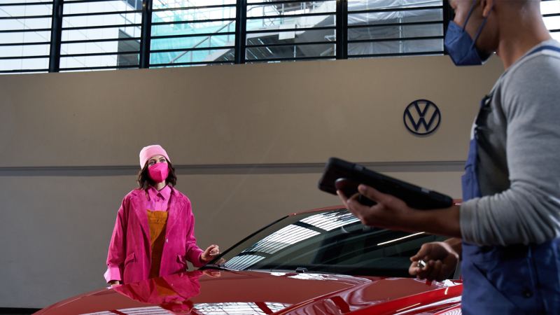 Eine Kundin steht an ihrem roten VW Auto und unterhält sich auf Distanz mit dem VW Servicemitarbeiter