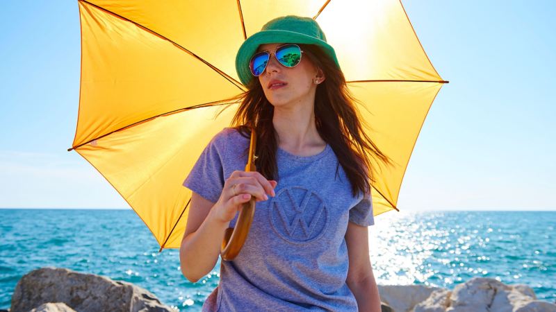 Eine Frau in Kleidung der Volkswagen Kollektion hält einen Schirm in der Hand, im Hintergrund das Meer