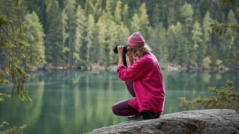 Eine Frau hockt auf einem Stein und fotografiert einen Bergsee