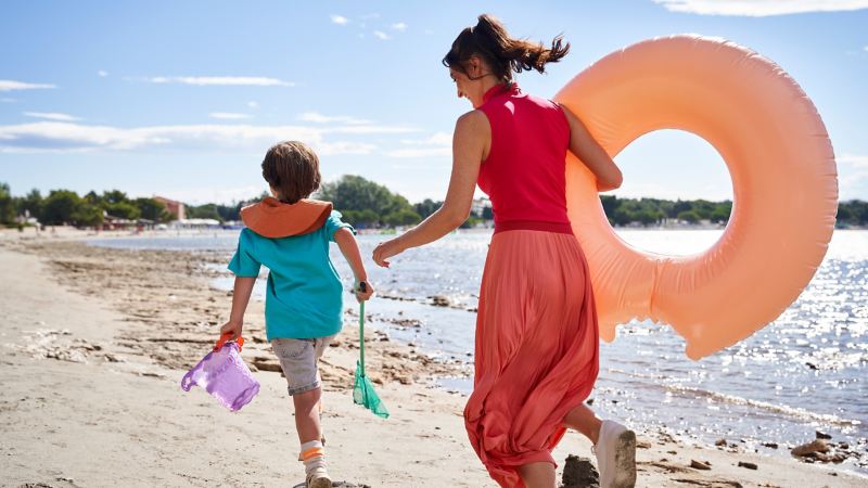 Eine Frau mit Schwimmring und ein Kind mit Kescher und Eimer laufen am Strand um die Wette