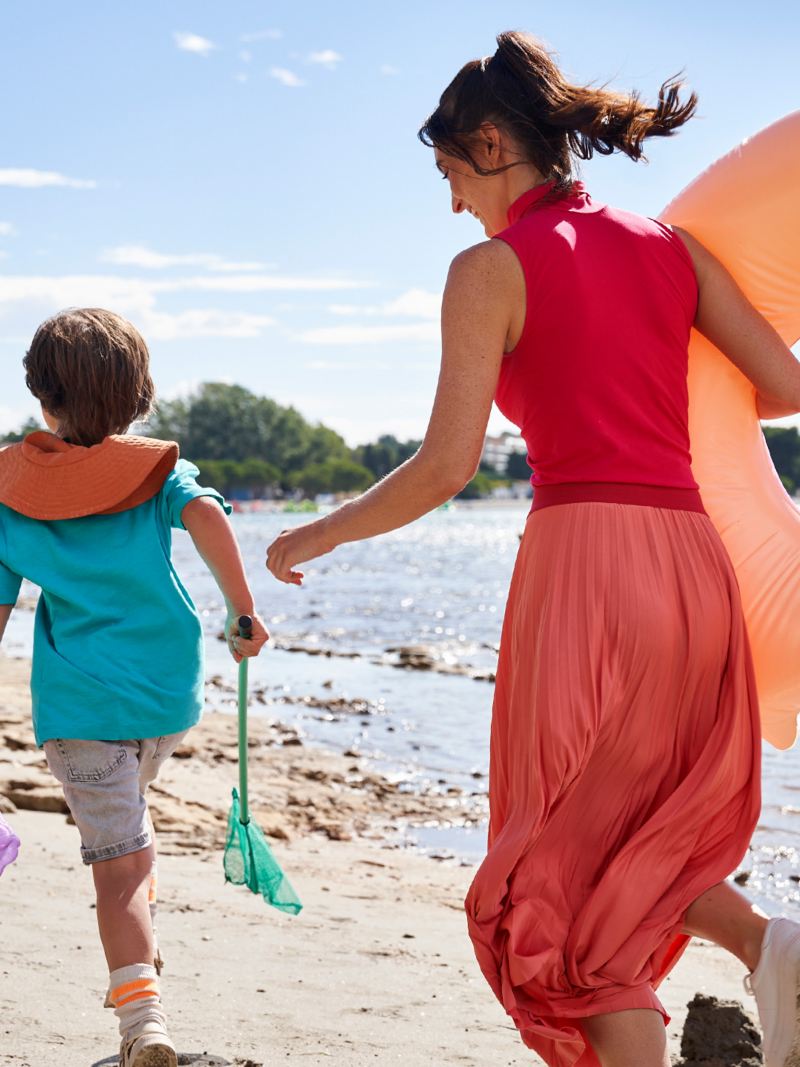 Eine Frau mit Schwimmring und ein Kind mit Kescher und Eimer laufen am Strand um die Wette