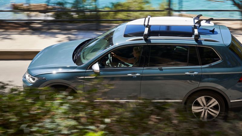 Der VW Tiguan Allspace fährt mit dem Surfbretthalter via Tragstäben auf dem Dach eine Straße entlang – VW Zubehör