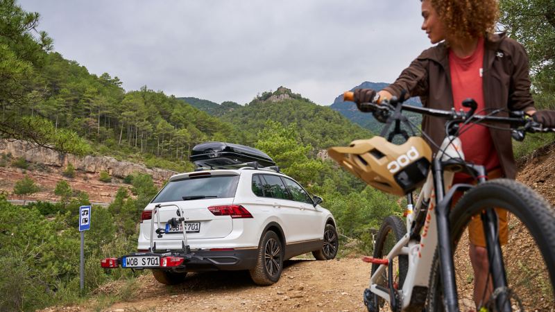 Femme avec un vélo, VW Tiguan avec coffre de toit et porte-vélos de la gamme d’accessoires VW en arrière-plan