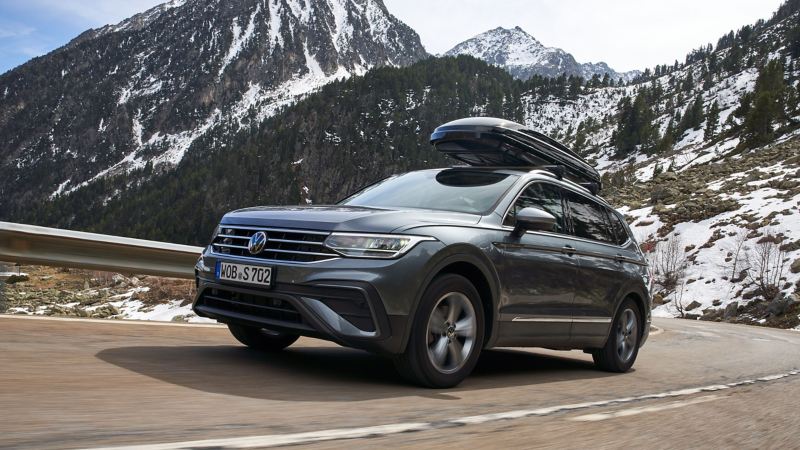 Un Volkswagen Tiguan équipé d’un coffre de toit Volkswagen Accessoires sur une route de campagne, montagnes en arrière-plan