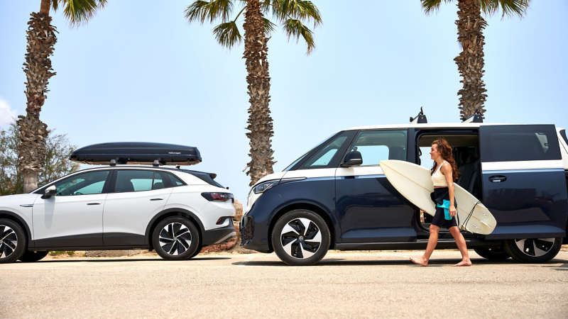 Una mujer con una tabla de surf pasa junto a un VW ID. Buzz, a su lado un VW ID.4