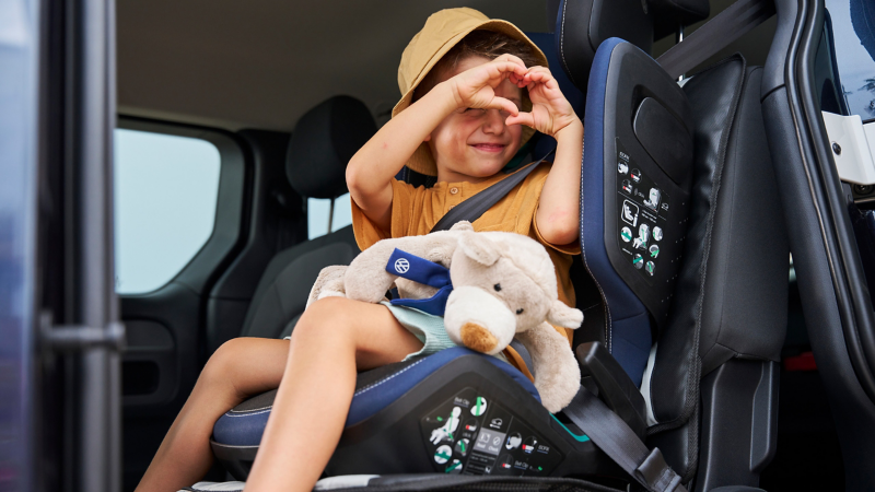 Boy in a VW Accessories child seat with cuddly toy on his lap forms a heart with his hands