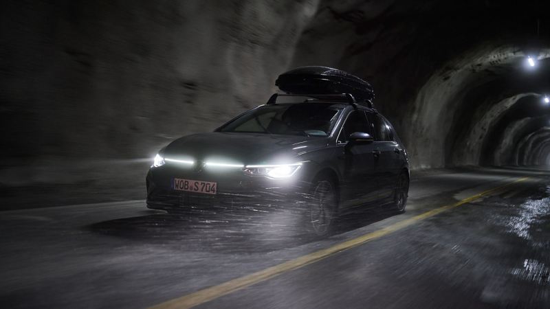 Un véhicule VW avec un coffre de toit roule dans un tunnel, phares allumés