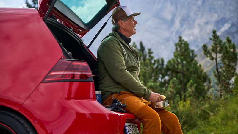 Man with a food container in his hand sits on luggage compartment lid of a VW Golf 7 and looks out into nature