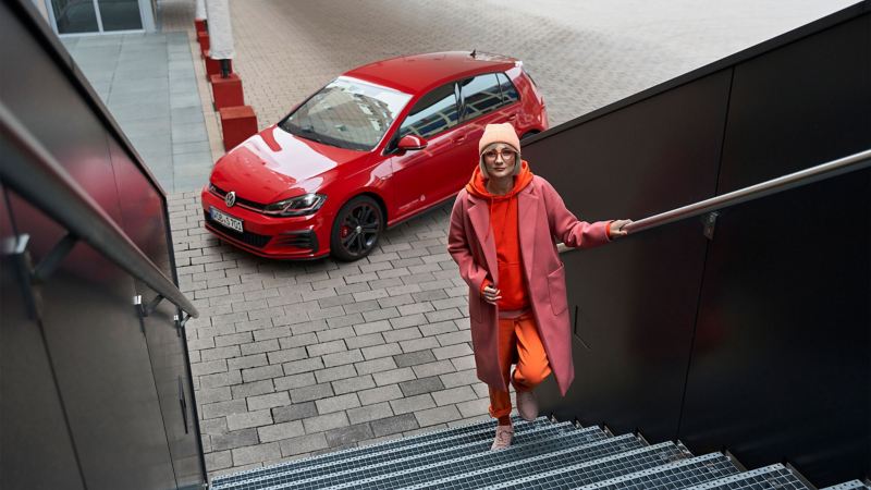 A woman walking up the stairs, in the background stands a VW Golf 7