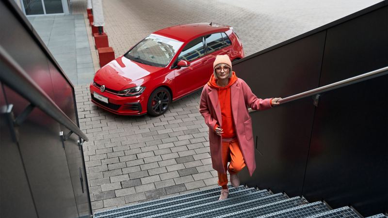 A woman walking up the stairs, in the background stands a VW Golf 7