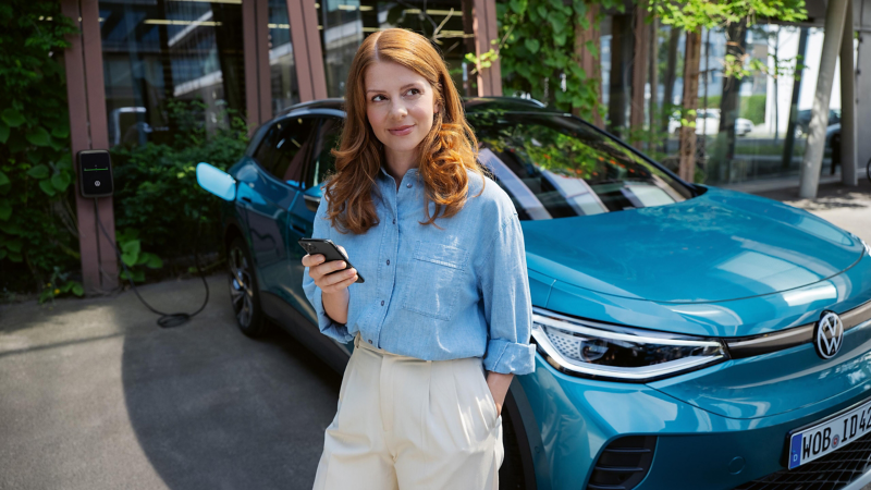 Frau mit Handy steht neben VW ID.4, der an ID. Charger Wallbox lädt