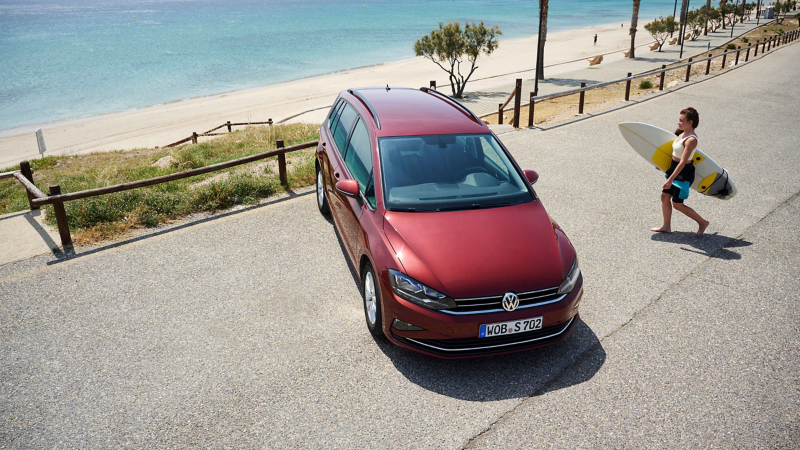 Frau mit Surfbrett neben ihrem VW Vorgängermodell am Strand