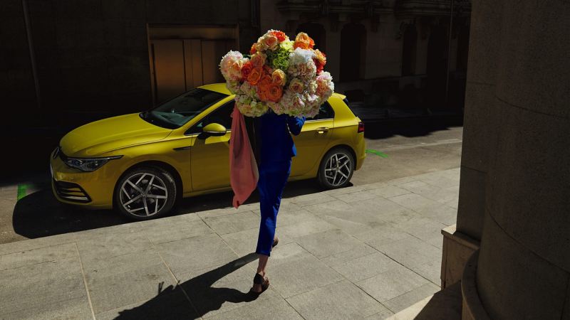 Mujer paseando con un ramo de flores al lado de un Volkswagen Golf amarillo