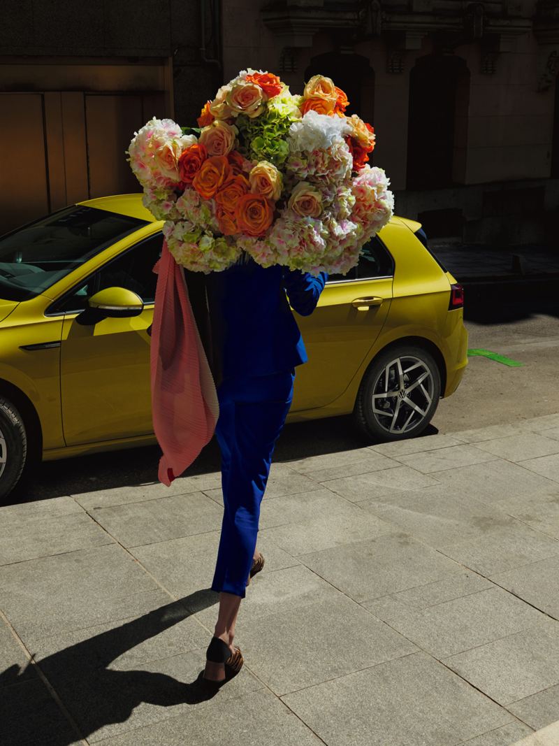 Una mujer vestida de azul con un gran ramo de flores se acerca a un VW Golf 8