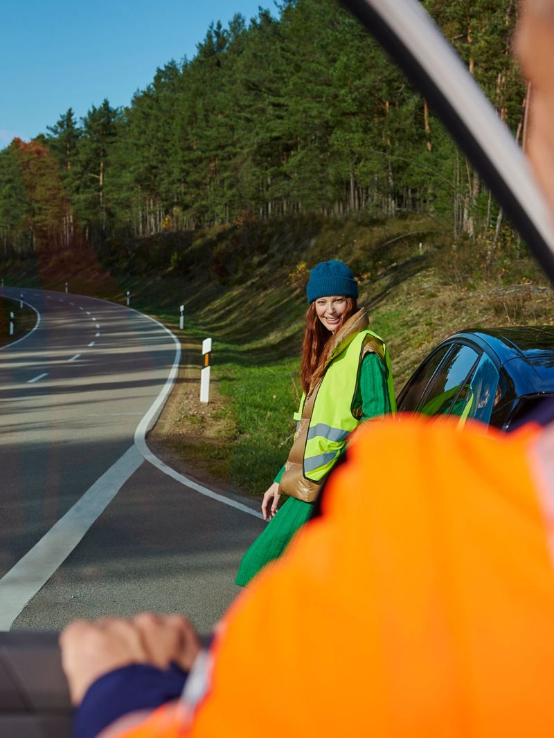 Ein VW Servicemitarbeiter der Pannenhilfe steigt aus seinem Auto, im Hintergrund lehnt eine Frau an ihrem VW Auto und wartet auf ihn 