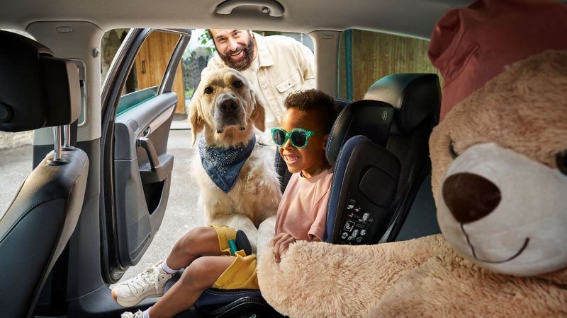 Mann und Hund schauen durch offene Tür auf Rückbank eines VW Autos zu Kind und Kuscheltier