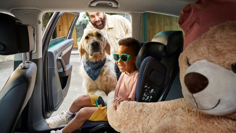 Un homme et un chien regardent à travers la porte ouverte un enfant avec une peluche sur la banquette arrière d’une voiture VW