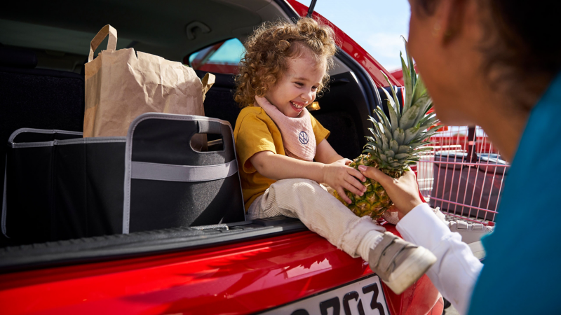 Frau gibt Ananas an Kind, das im Kofferraum eines VW Polo 5 sitzt