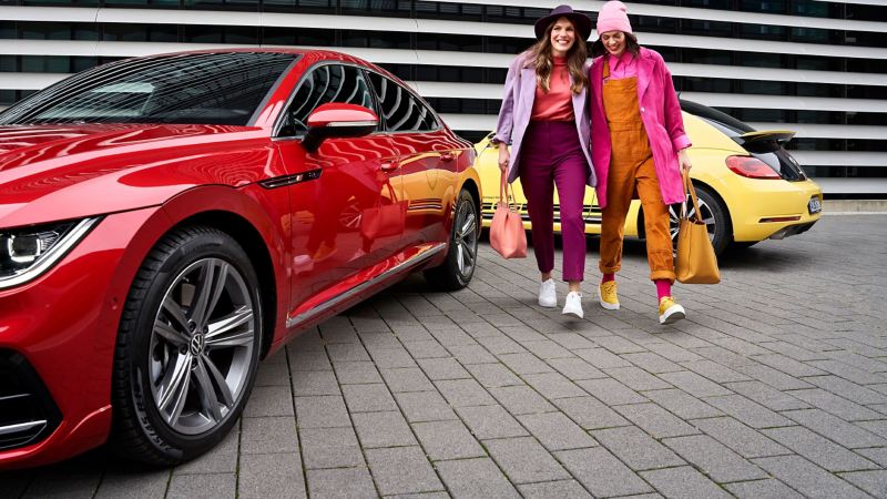 Zwei lächelnde Frauen vor ihren VW Autos – ein roter Arteon und ein gelber VW Käfer