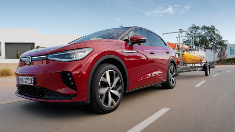 Un SUV électrique rouge Volkswagen ID.5 tirant une remorque avec deux kayaks jaunes, roulant sur une route goudronnée dans un environnement ensoleillé et résidentiel.