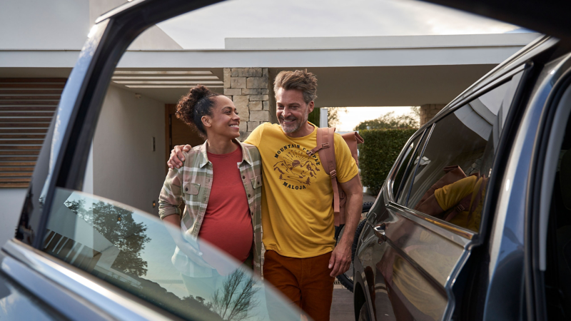 Man brings pregnant woman to a VW car, in the foreground the open passenger door