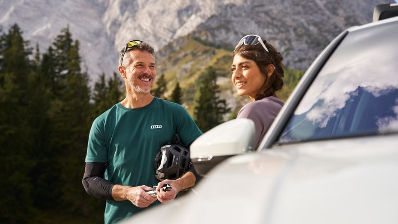 Mann und Frau mit Sonnenbrillen auf dem Kopf stehen neben VW Tiguan 2, im Hintergrund idyllische Bergkulisse