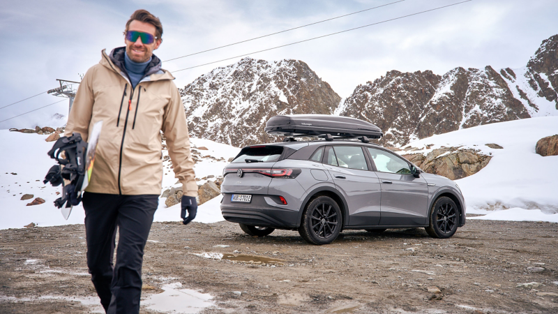 VW ID.4 con box da tetto in un paesaggio invernale innevato, in primo piano un uomo con uno snowboard.