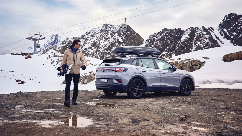 VW ID.4 Pro con box da tetto in un paesaggio invernale innevato, in primo piano un uomo con uno snowboard.