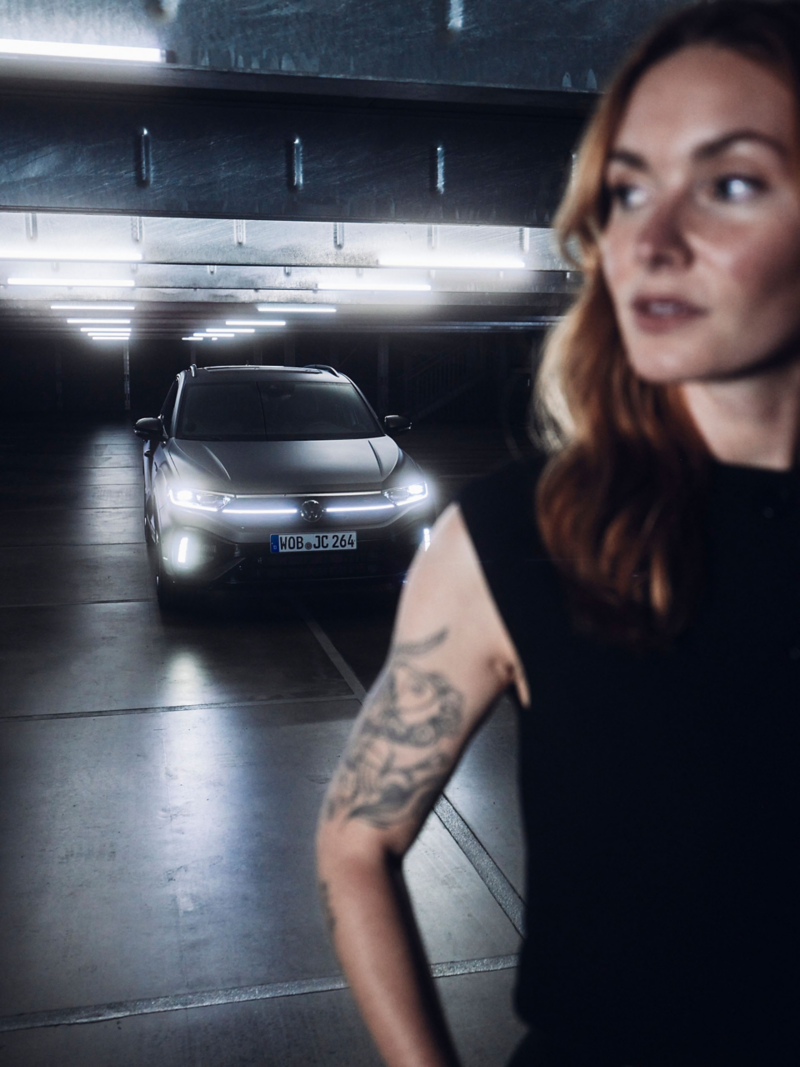 Front view of VW T-Roc R with illuminated headlights, woman standing in the foreground