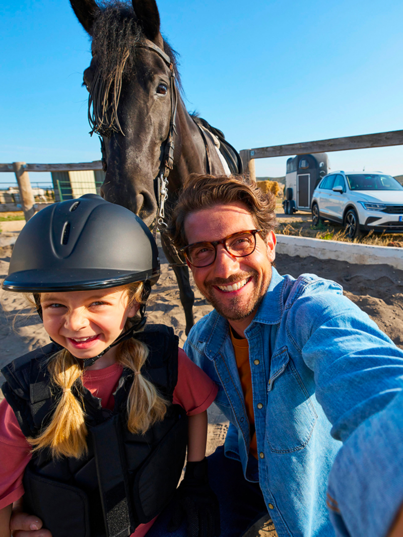 Un padre scatta un selfie con la figlia davanti al cavallo, sullo sfondo VW Tiguan con rimorchio per cavalli agganciato.