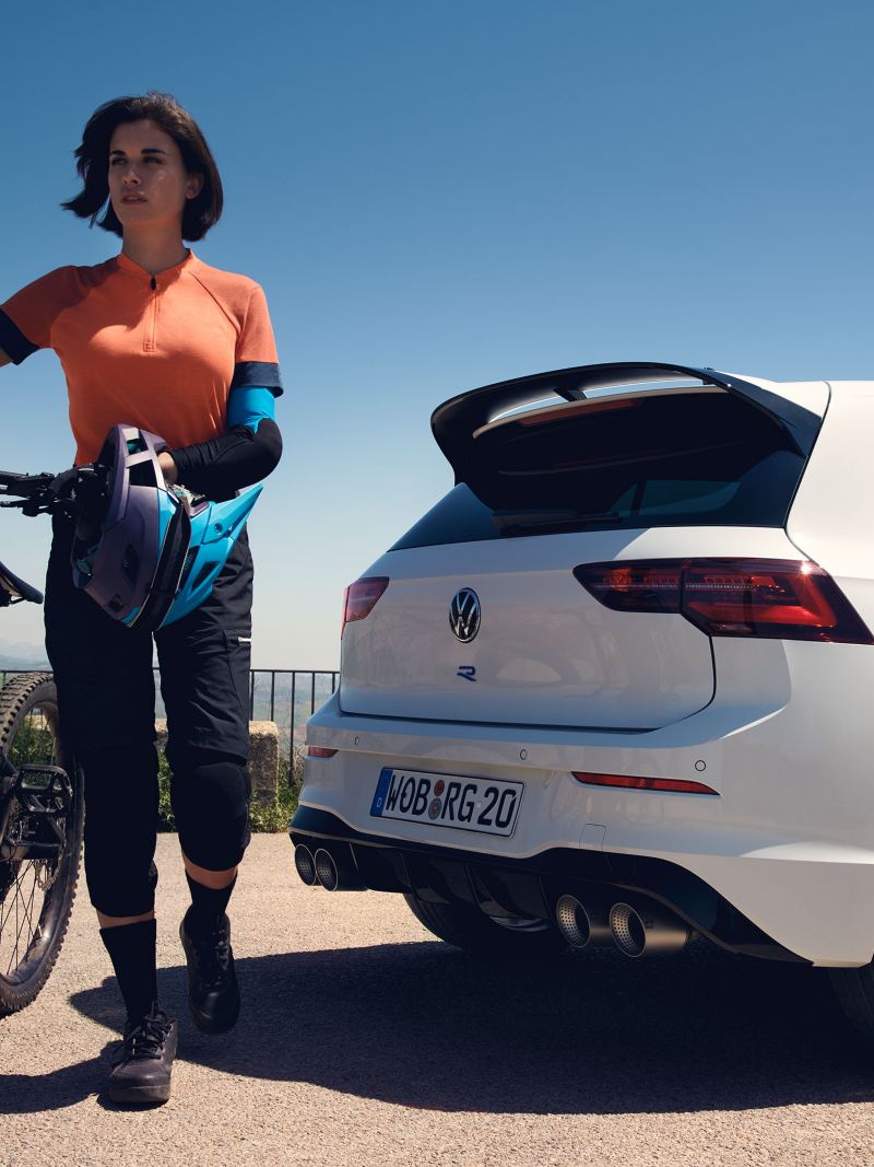 Una donna in piedi con la sua mountain bike accanto alla sua Golf R bianca «20 Years» di VWR