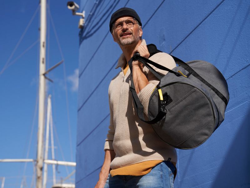 Ein Mann mit Fischermütze trägt eine Reisetasche von VW Zubehör, im Hintergrund ein Segelboot