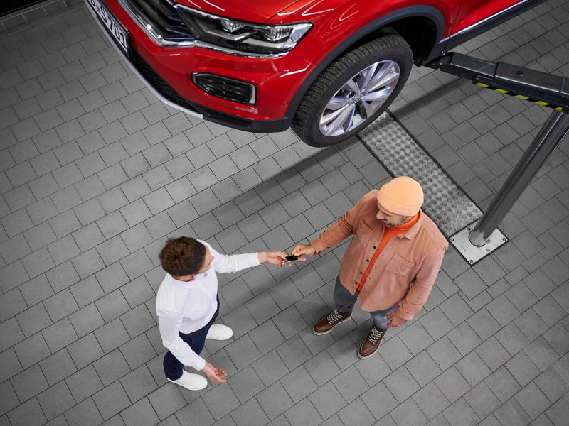 Ein Kunde überreicht seinen Autoschlüssel an eine Servicemitarbeiterin, auf der Hebebühne ist ein roter VW