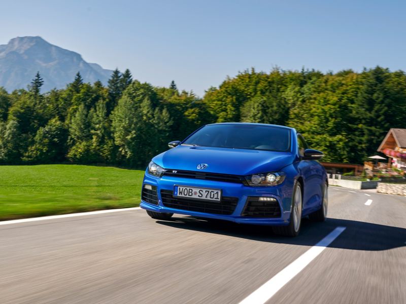 Blauer VW Scirocco 3 fährt auf einer Landstraße, im Hintergrund Berge