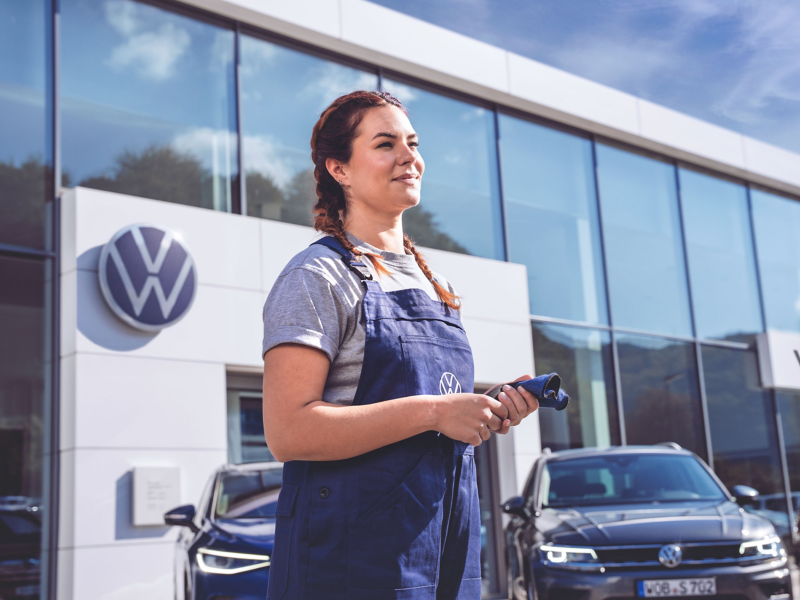 VW Servicemitarbeiterin vor einem VW Autohaus hält Arbeitshandschuhe in der Hand