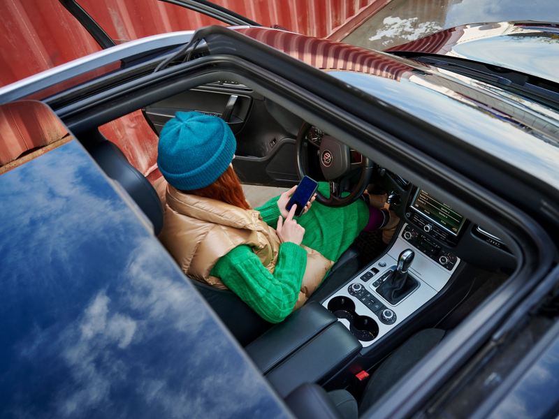 Eine Frau sitzt im geparkten VW Touareg mit offener Fahrertür und bedient ihr Smartphone – VW Zubehör