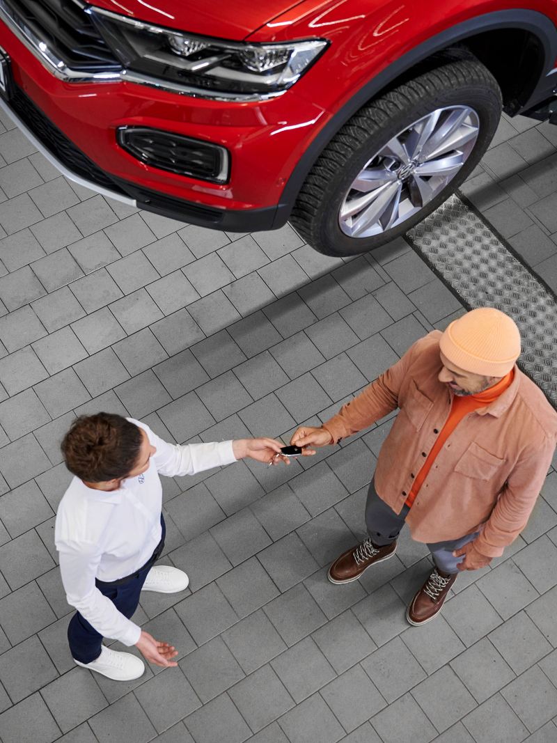 A customer hands his car key to a service employee, on the lift is a red VW