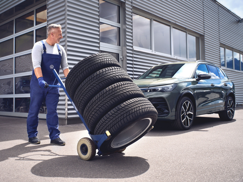 Addetto del servizio Volkswagen che spinge un carrello con quattro ruote impilate, sullo sfondo un VW Tiguan davanti all’officina.